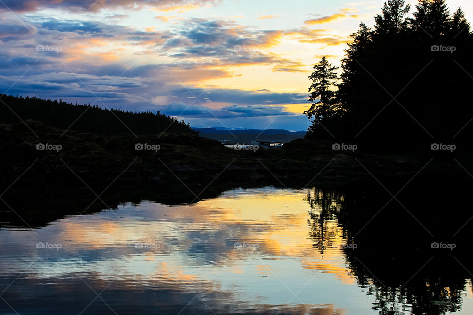 A silhouette of a small island partially covered with trees in an ocean inlet. The golden setting sun & darkening sky created beautiful shades of blue, purple, orange, yellow & pink in the sky & this was perfectly reflected in the still calm waters.