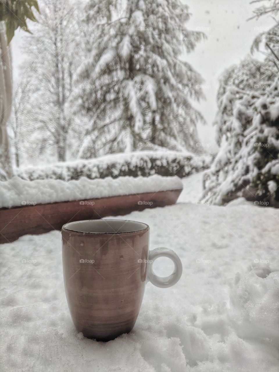View of the snowfall over the cup of hot drink and snow-covered trees in morning winter.