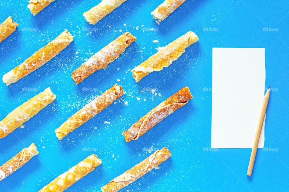 Dynamic composition with homemade cakes. Delicious waffle tubes with powdered sugar on a bright blue background. Delicious sweet snack, top with a torn white sheet of paper to record the recipe and a pencil. Children's treat. 