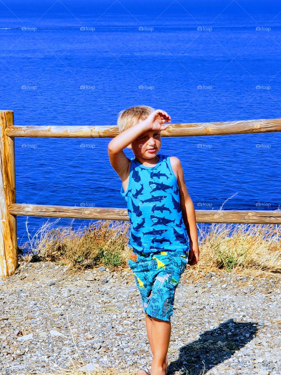 a little boy by the sea is blocking the sun with his hand