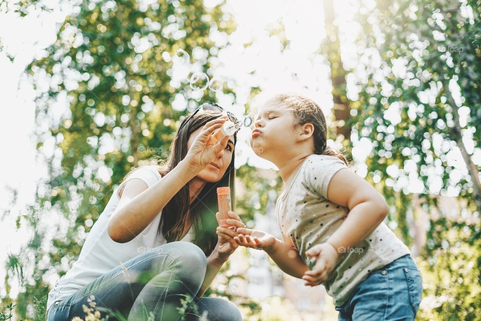 Cute toddler girl blows on soap bubbles with his mother in sunny summer day