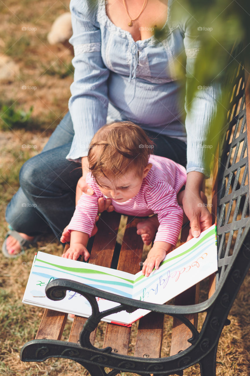 Mother and little baby sitting on park bench and reading book