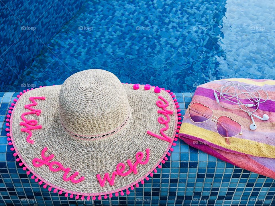 Summer hat with the words “wish you were here”,sunglasses, headphones and a pink towel on the edge of a pool with turquoise water