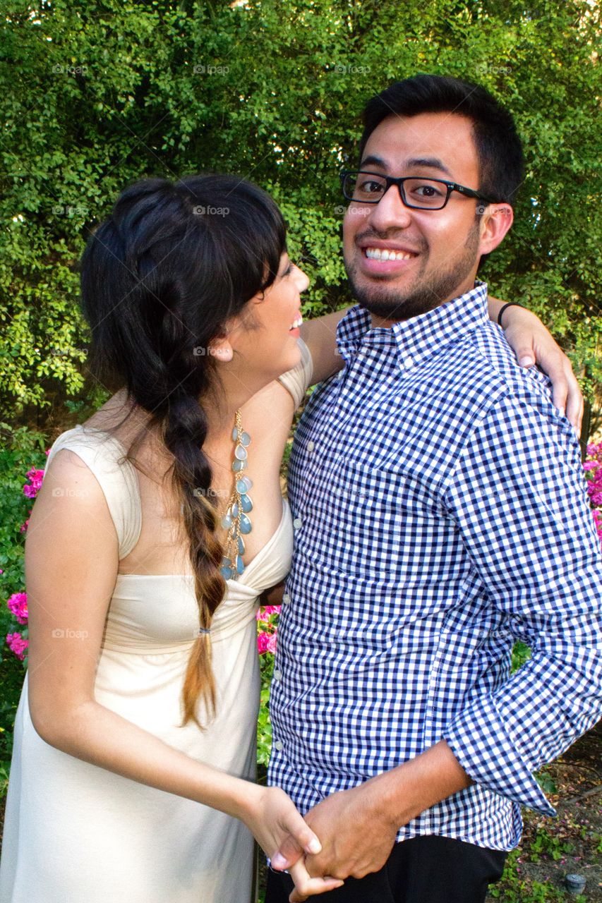 Happy young couple laughing and holding hands in front of tall green bushes and pink flowers in a garden