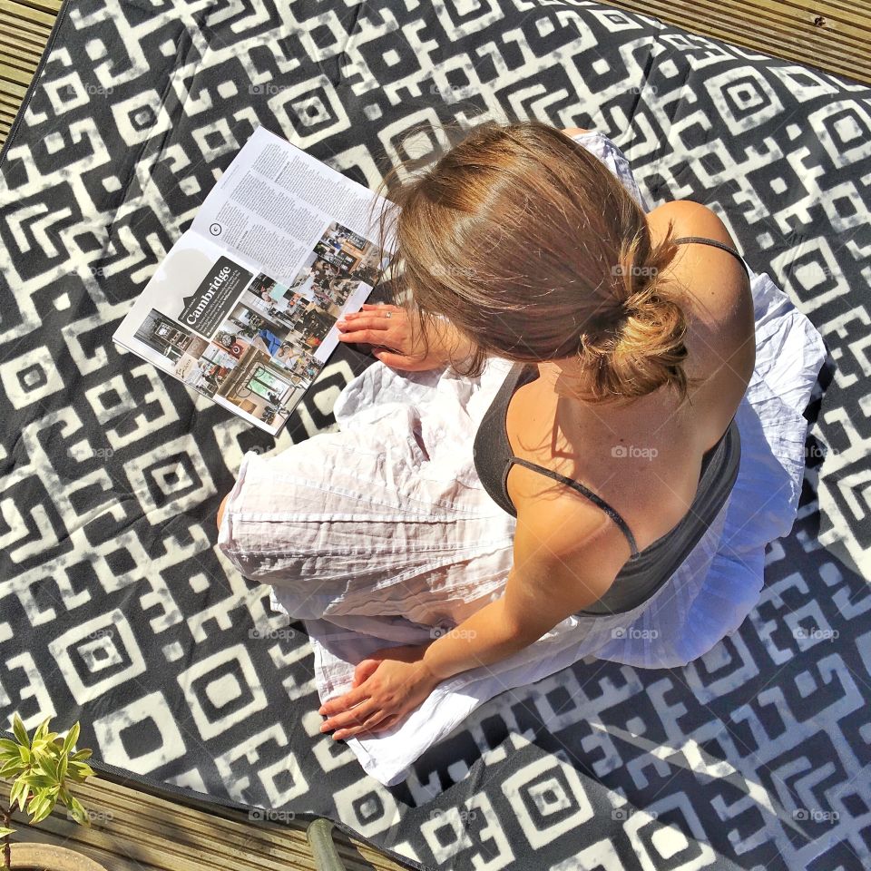 Young woman from above reading magazine sitting on the blanket cross legged in the sun. 