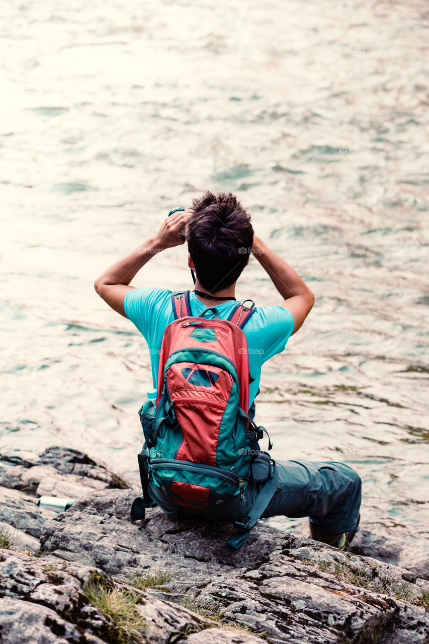 Young tourist with backpack looks through a binoculars, sits on a rock over a river. Boy spends a vacation on wandering with backpack, he is wearing sports summer clothes