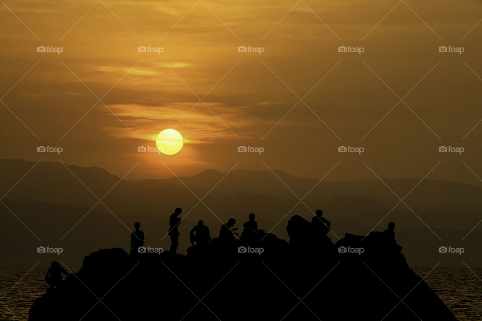 Silhouette of tourists Fishing on the rocks in sea and Golden light of sunrise behind the mountains.