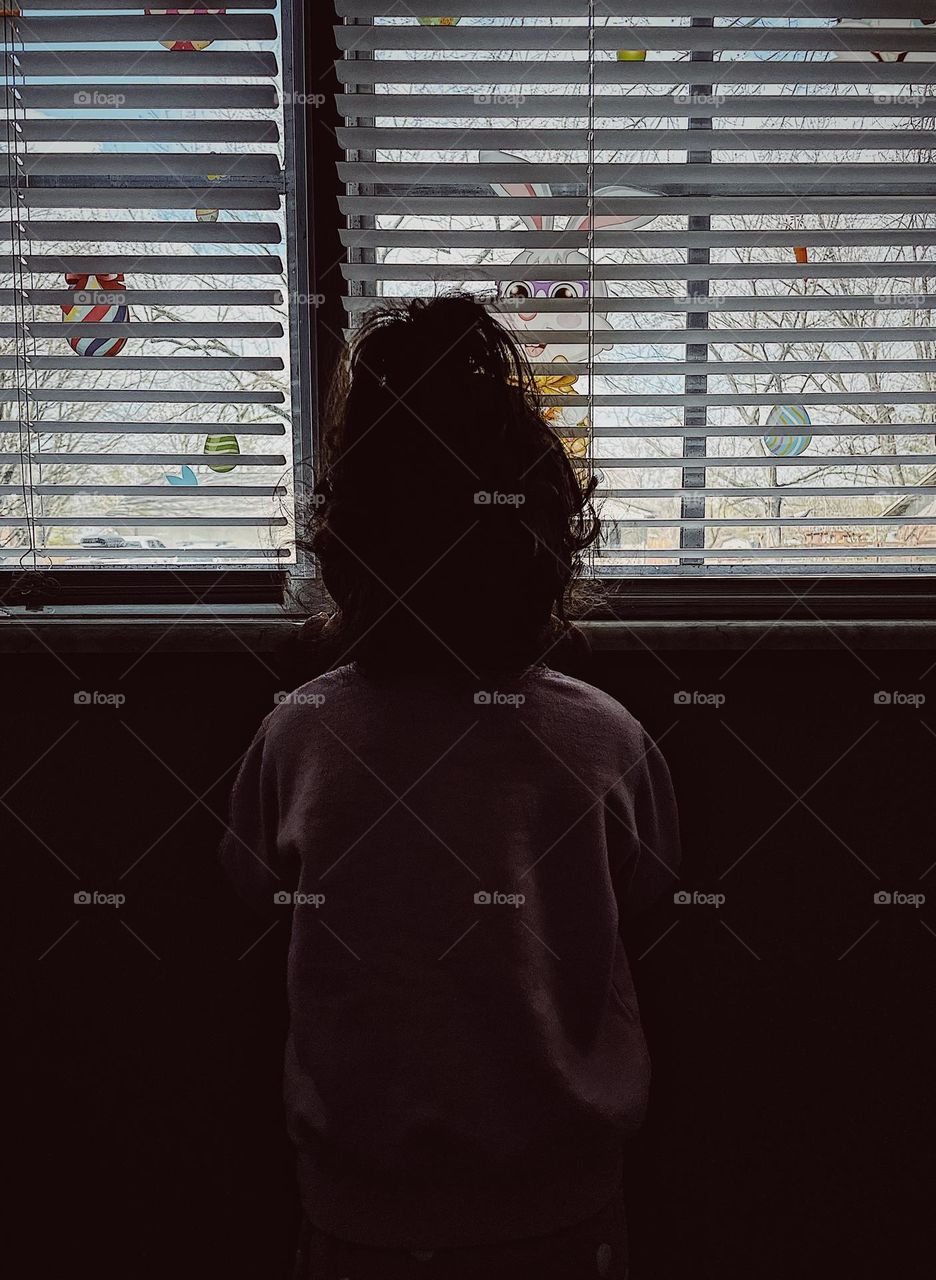 Little girl standing at window looking out, little girl in the shadows looking towards the light, windows of the home decorated with Easter decor, toddler girl curious about the neighborhood, little girl watches cars go by on the street