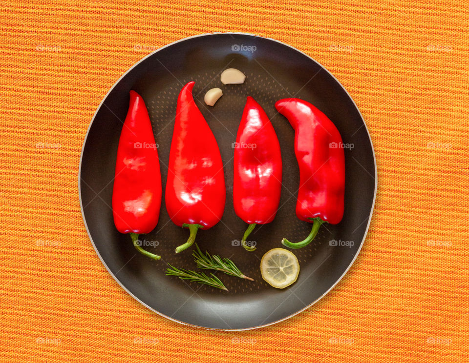 Baking dish with red sweet bell peppers with rosemary sprigs, lemon wedges and garlic on a bright orange textured textile background. Ingredients for cooking grilled peppers