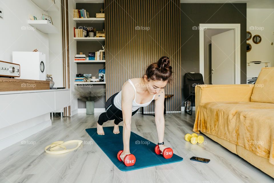 Millennial woman dressed in a white tank top and black leggins, working out at home, using weights and elastic band.