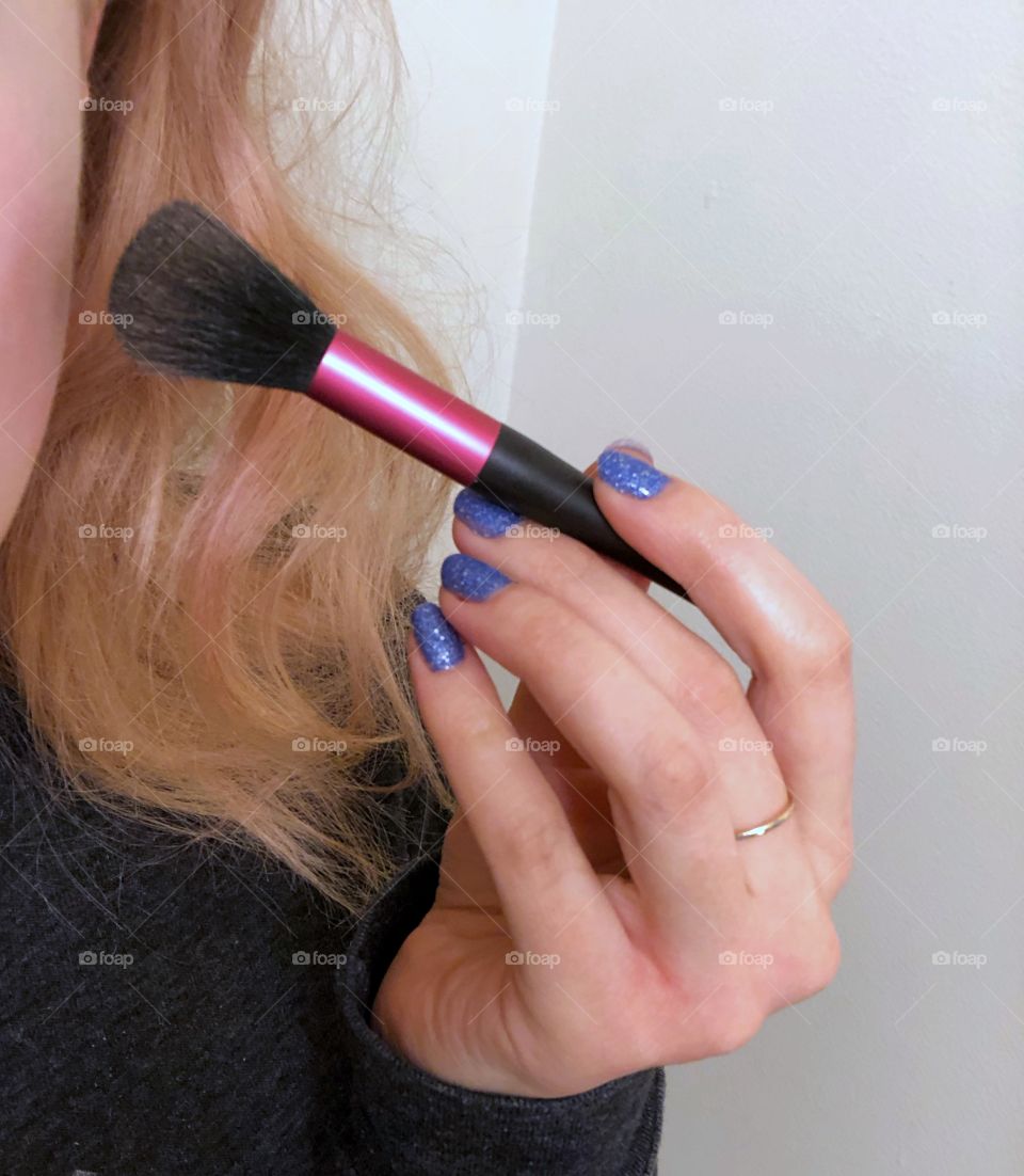 A woman’s hand with bright blue fingernails, holding a make up brush and applying blush to her cheek