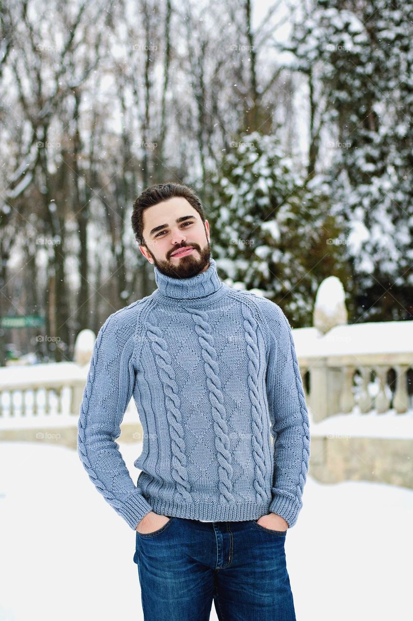 Portrait of handsome young smiling man in blue sweater in snowy winter park. Beard. Bearded guy, man.