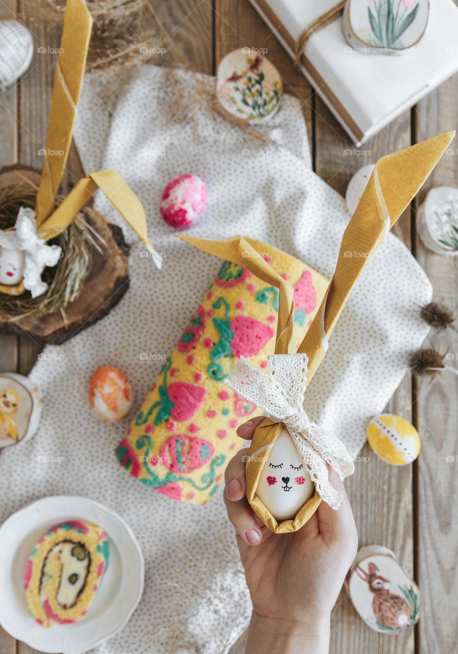 Overhead photo of hand holding easter egg decorated as a bunny over wooden table with easter foods and cake