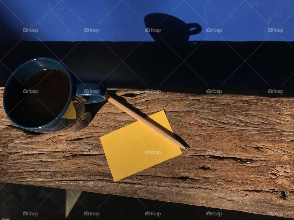 hot black coffee’s put on natural wood bench with blank yellow note paper and brown pencil, and its shadow reflects on navy blue wall in present morning