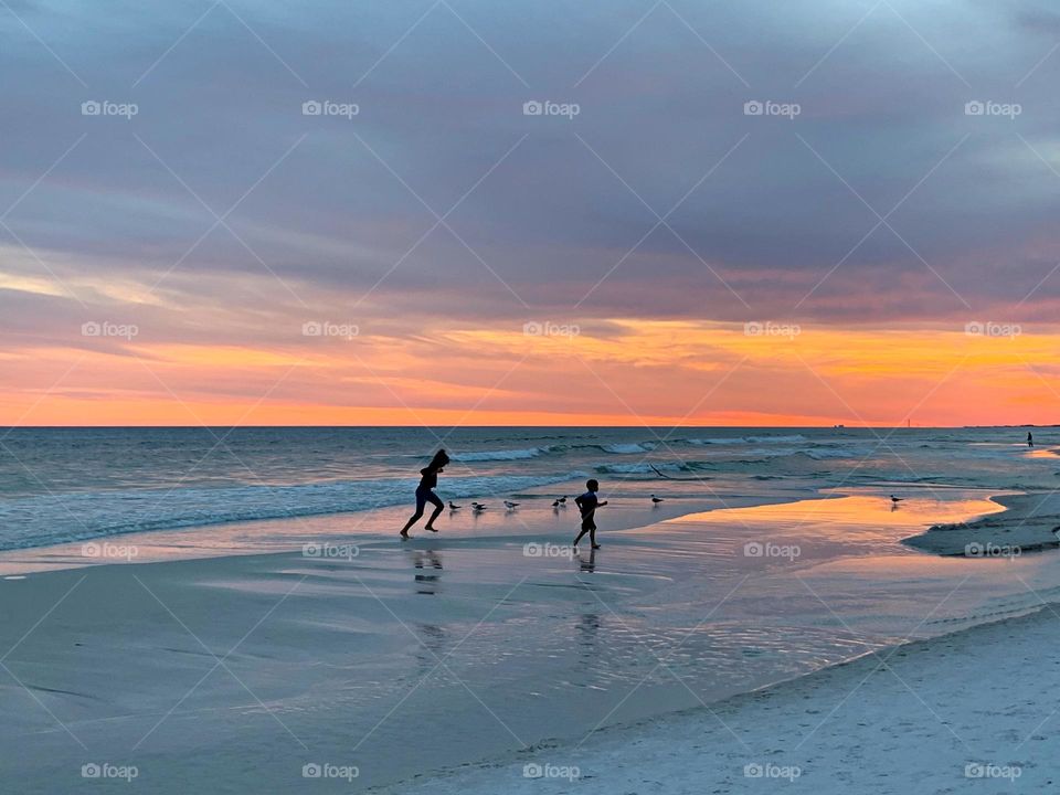 A beach at sunset, characterized by the warm hues of the sky reflecting on the wet sand and ocean. A silhouetted parent and a child, are seen on the wet sand near the water's edge, playing.