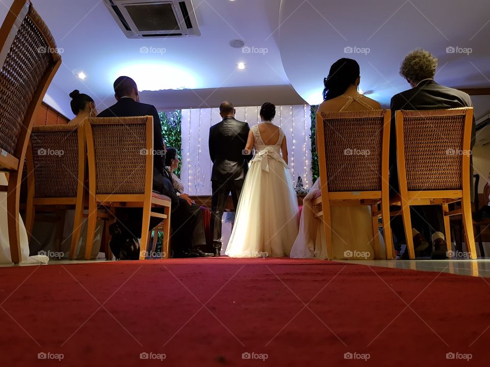 Cerimônia de casamento. Noivos em pé entre padrinhos sentados, todos de costas. Foto tirada a partir do solo.