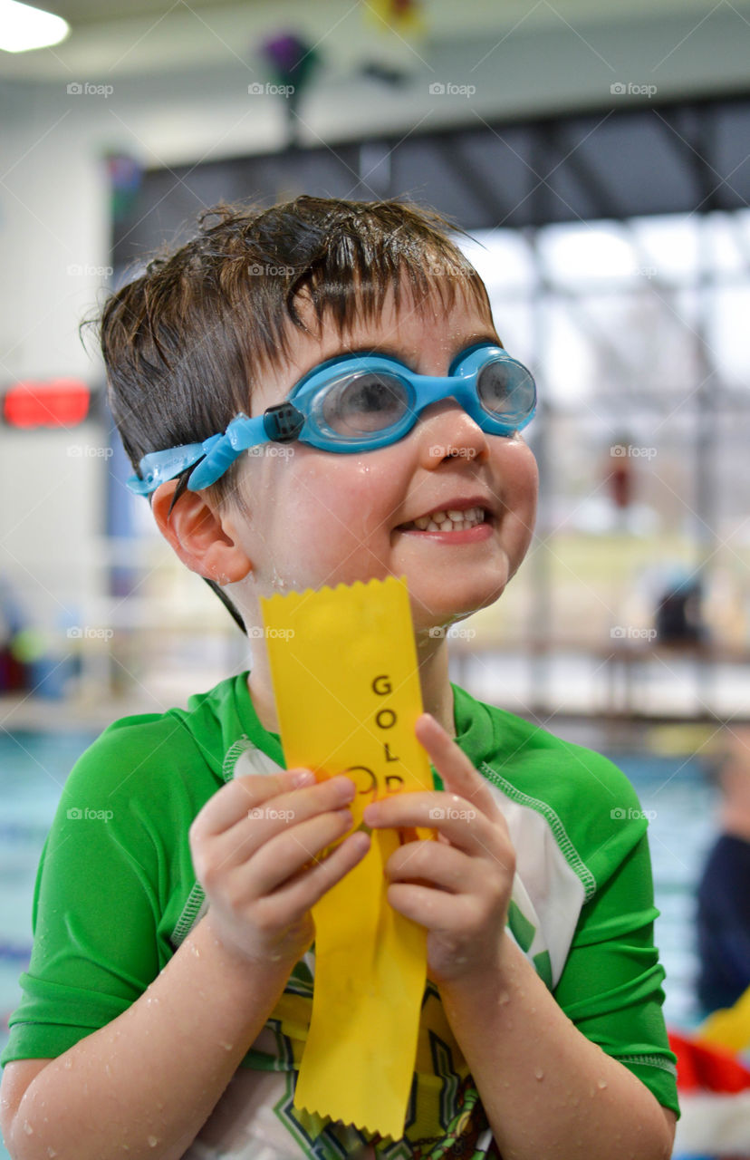 Young toddler boy wearing swimming goggles indoors smiling and holding an award ribbon after a swim lesson