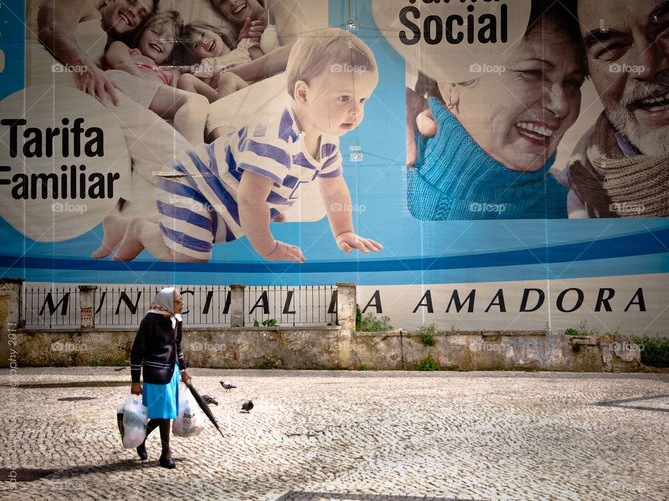 Black old woman walking and carrying plastic bags in a street of the suburbs. Behind her there's a giant outdoor announcing social measures by the local authorities.