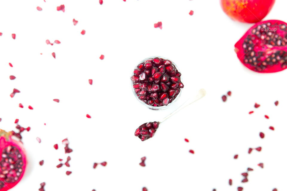 Fresh pomegranates on a white background. Lay flat image with glass bowl filled with fresh red pomegranate seeds, whole and cut pomegranates on the edges with scattered pomegranate seeds on the background. Fresh summer fruit encourages a healthy life