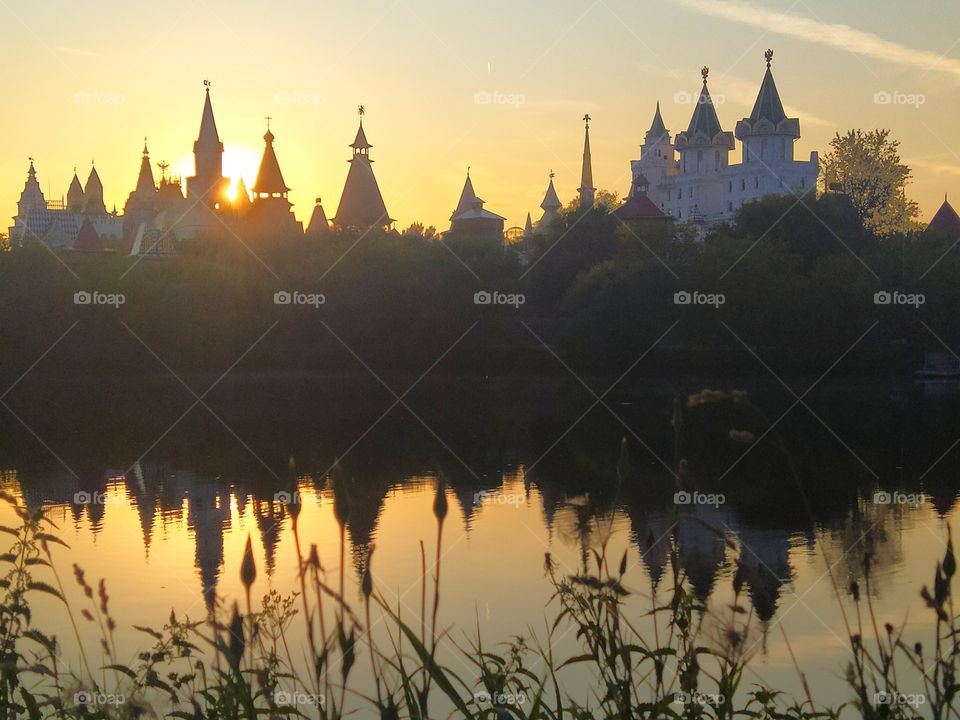 River.  The domes of the towers are visible among the trees across the river.  The sun sets behind one dome of the tower.  All surrounding objects are reflected in the water