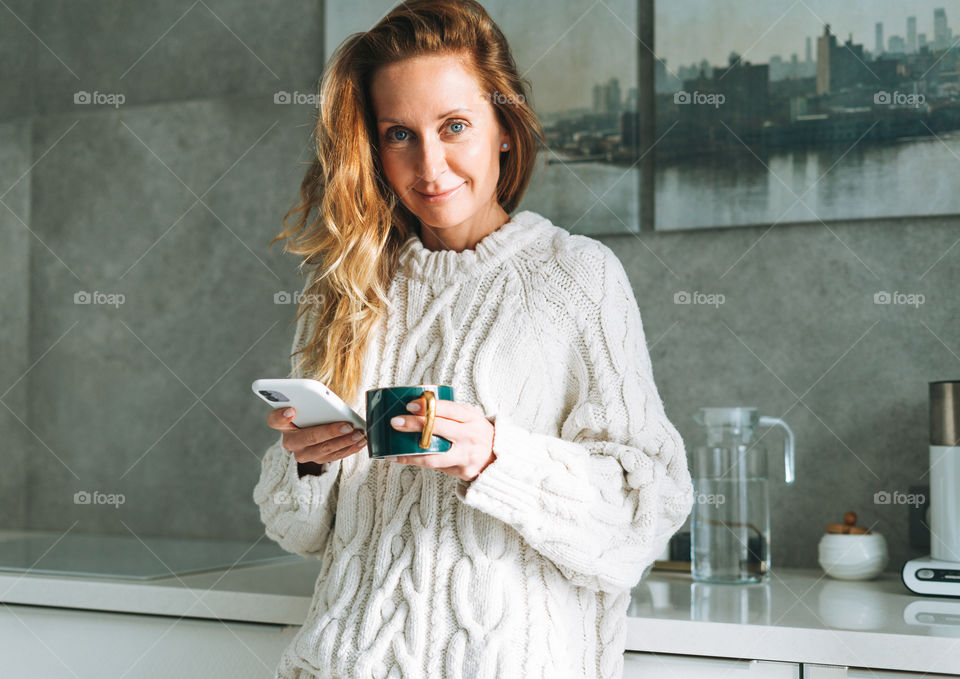 Young woman with long hair in knitted white sweater using mobile phone with cup of coffee in kitchen at home