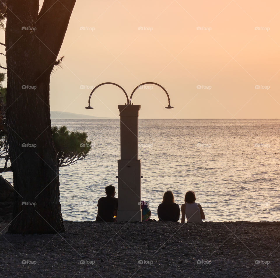 Silhouettes of young people sitting on a pebble beach in the evening, admiring the sunset.  Croatia, Dalmatia, Brela, sunset in the sea on the beach of Punta Rata