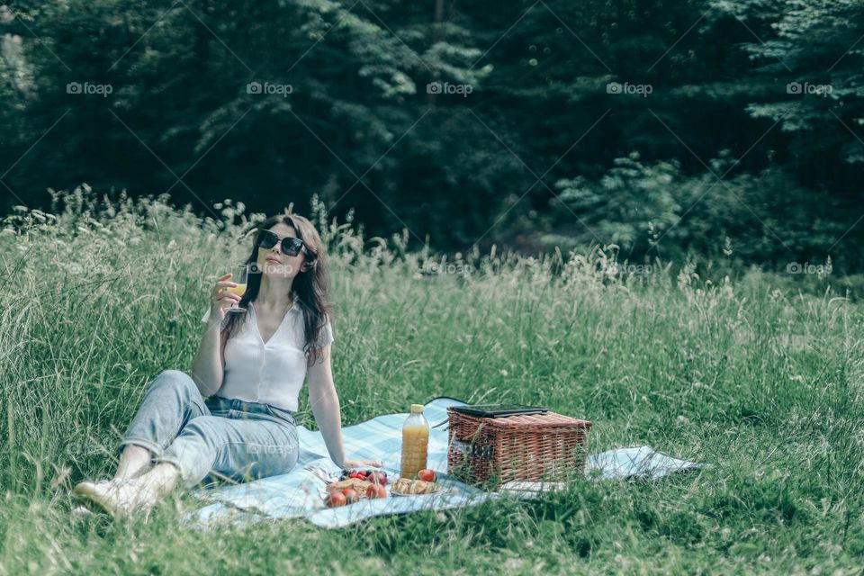 One young beautiful caucasian girl in sunglasses sits on a bedspread with a wicker basket, spread out food, drinks and holds a glass of orange juice in her hands looks to the side enjoying her vacation, close-up side view.