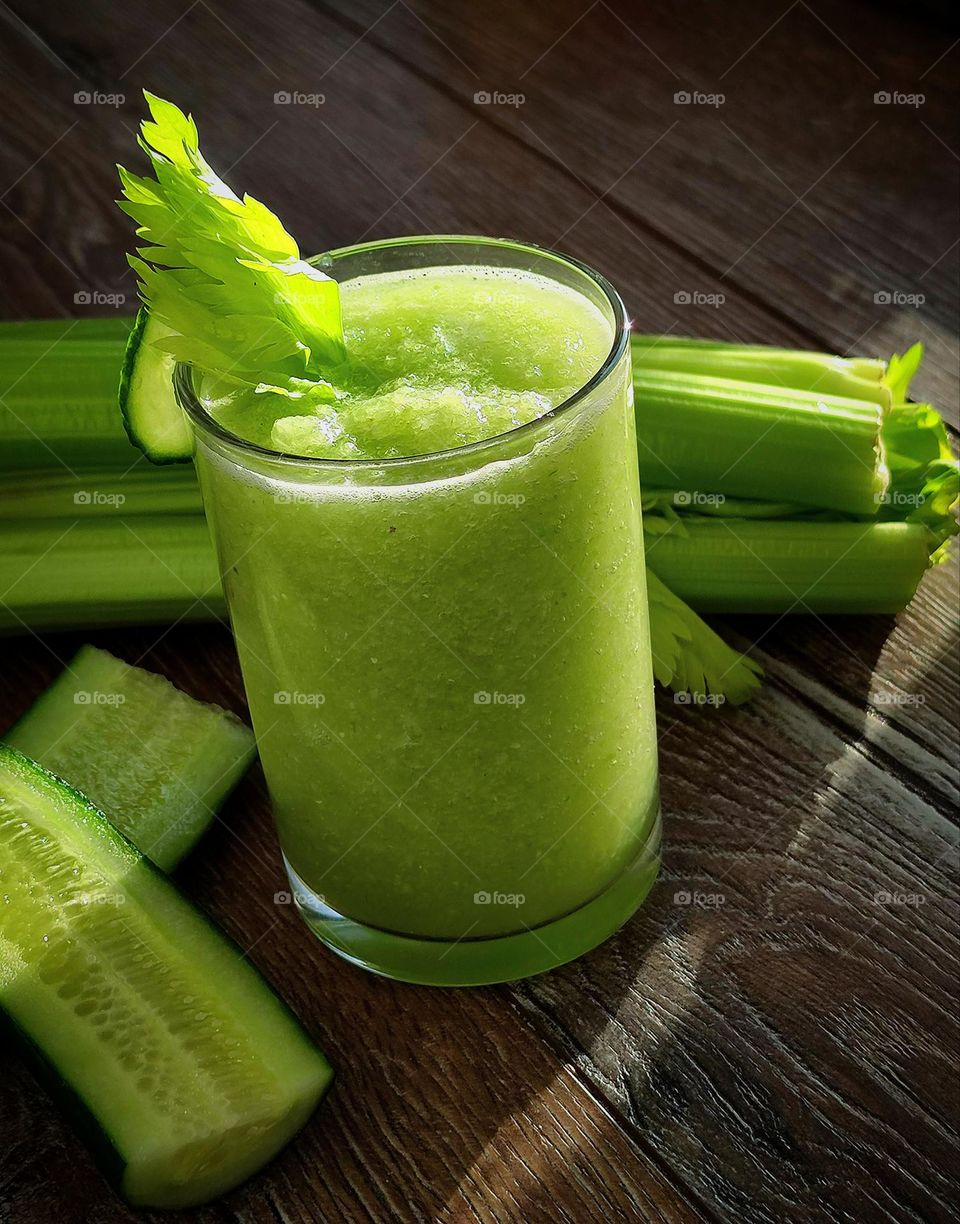 On a wooden surface is a glass of green smoothie garnished with a celery leaf. Nearby are the ingredients: a cucumber cut in half and a stalk of celery. The rays of the sun fall on the composition