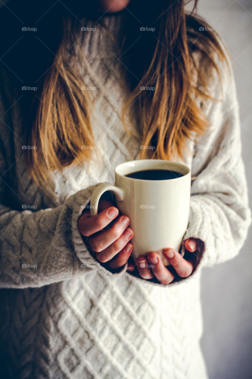 A blonde hair girl in a cosy, knitted sweater holding a cup of hot coffee in her hands
