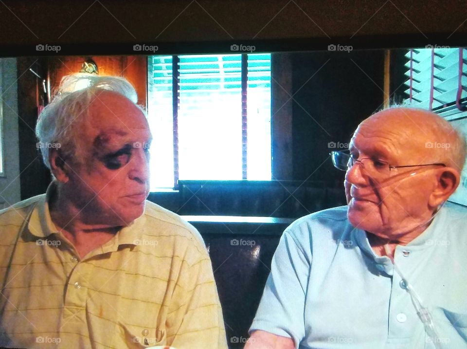 Older senior men sitting close in a table booth, just looking at each other. One older man has a black eye & his old friend is breathing with use of oxygen with use of oxygen tank & tubes.