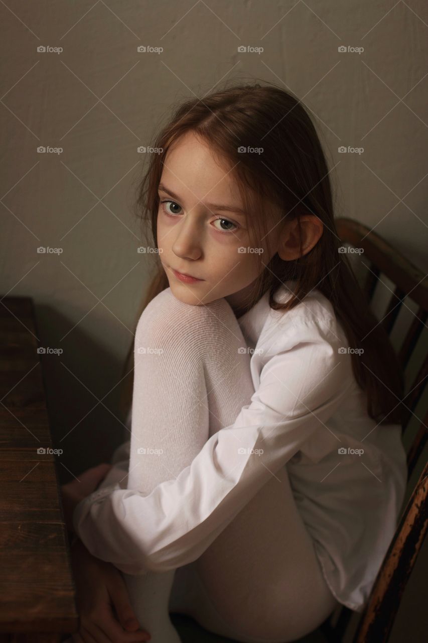 Little girl in a white shirt with long hair sits in the kitchen pulling her knee to her chin