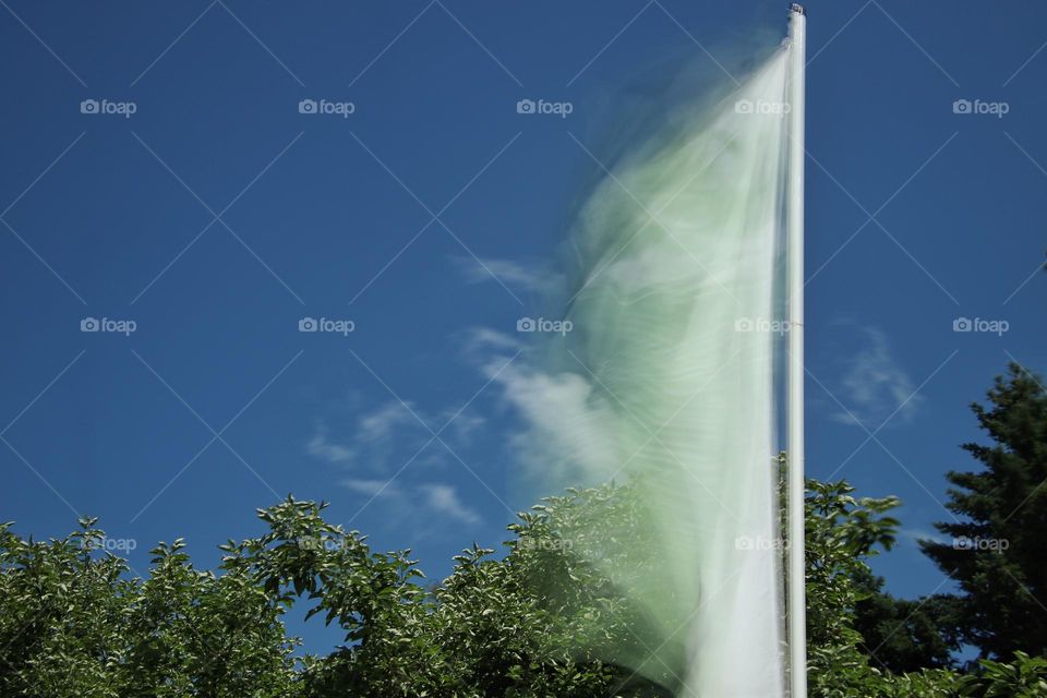 Long exposure of a waving white and green flag against a blue sky and trees 