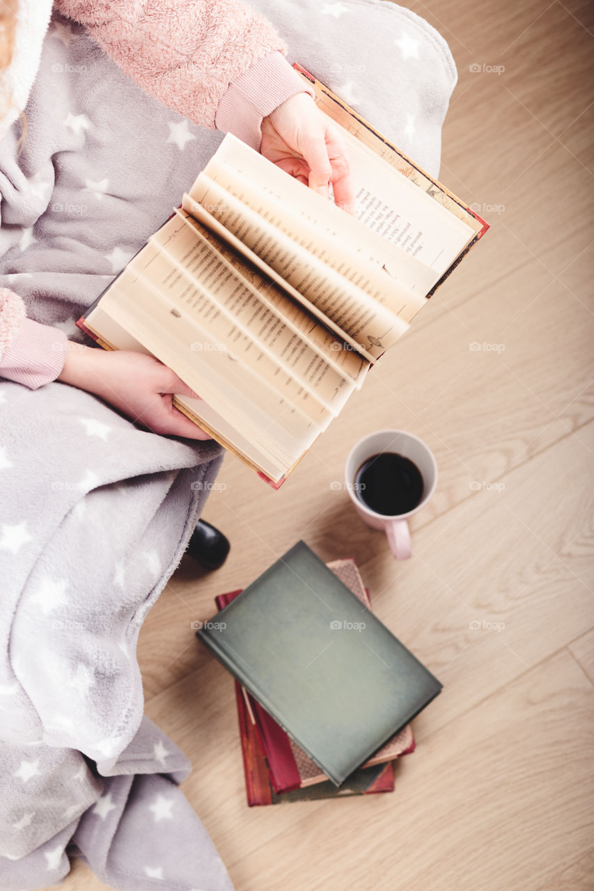 Girl enjoying the reading a book and drinking coffee at home. Young woman sitting on a chair, wrapped in blanket, holding book, relaxing at home. Portrait orientation. View from above