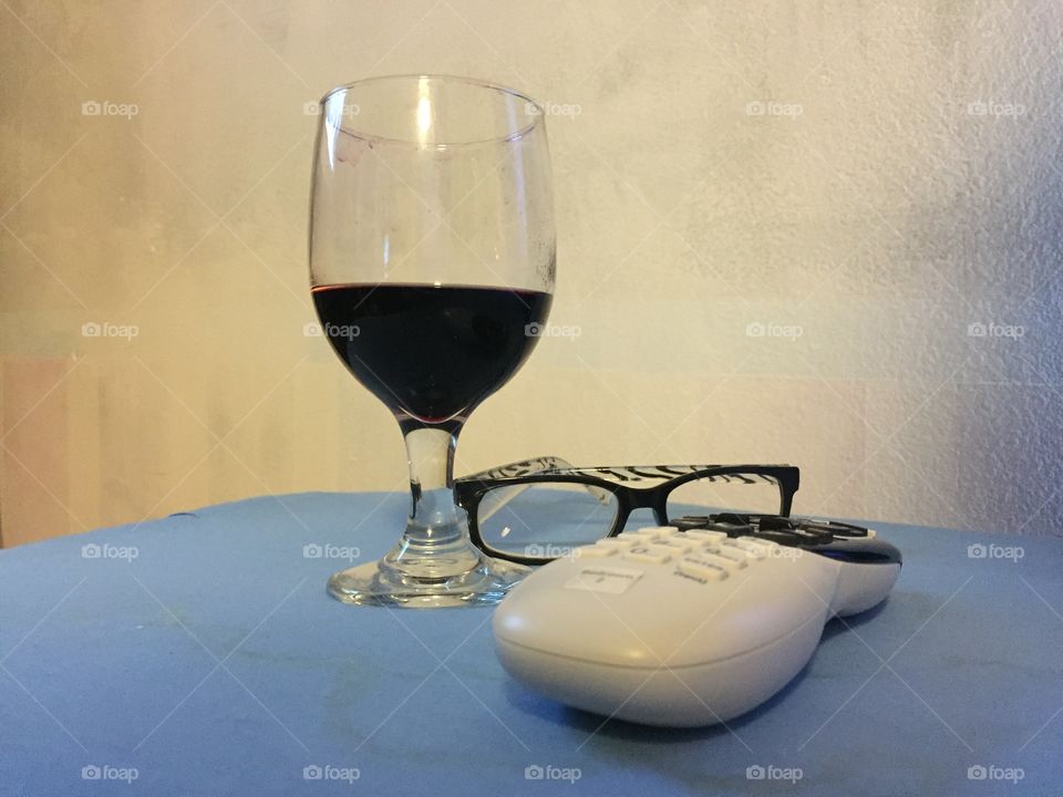 A glass of red wine a pair a reading glasses and a television remote control 