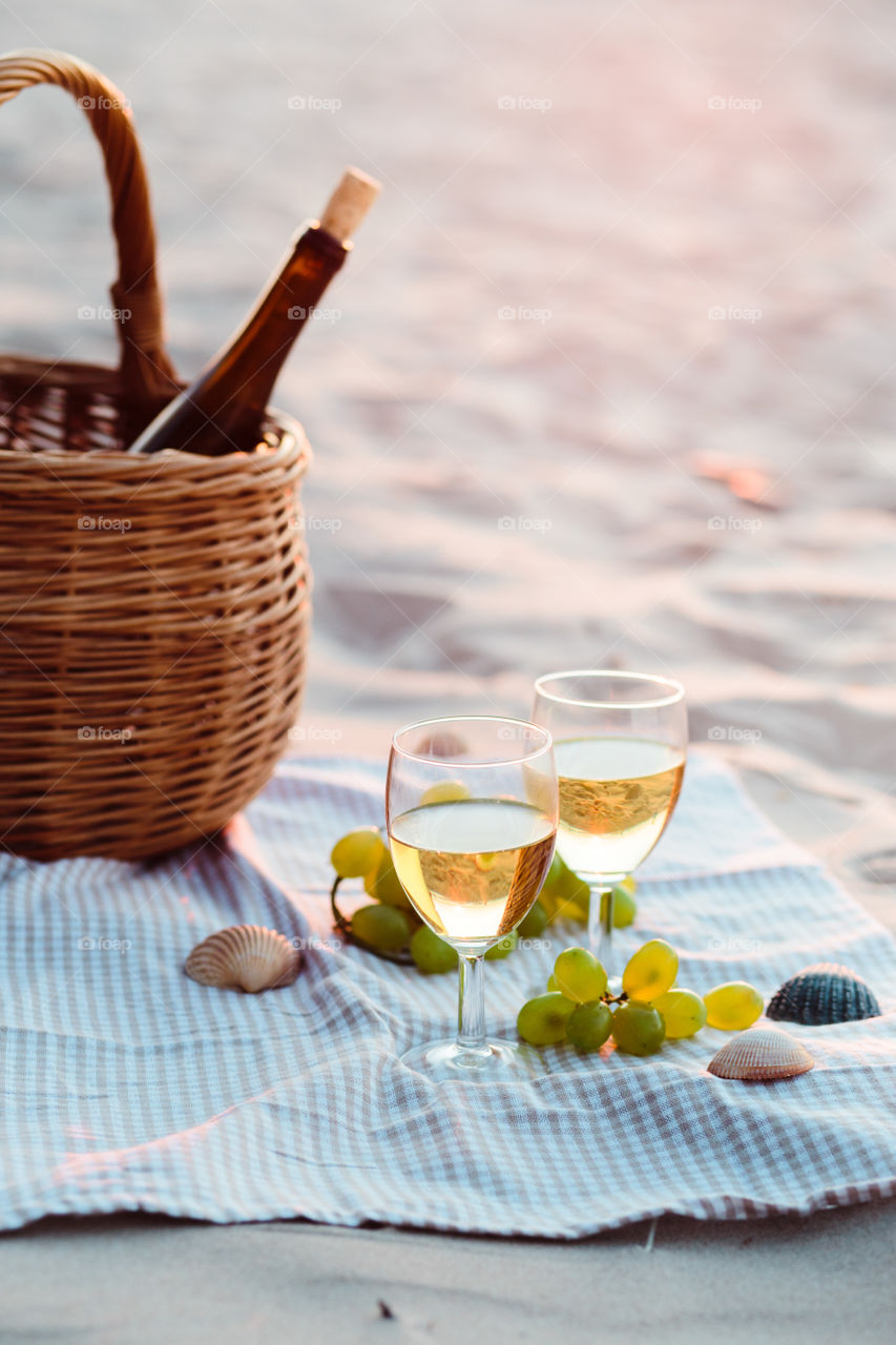 Two wine glasses with white wine standing on sand, on beach, beside grapes and wicker basket with bottle of wine. Sea waves in the background