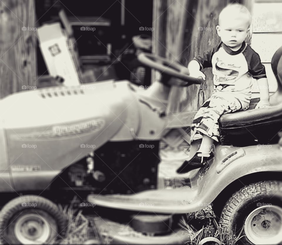 Classically cited monochrome moment captured of country baby boy driving lawnmower on summer day in yard.