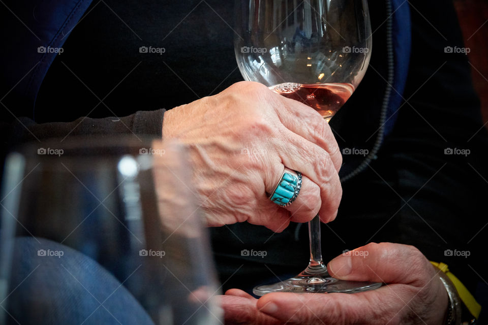 A wealthy old woman’s hands with a large blue ring holds a wine glass in conversation. Another glass is out of focus in the foreground indicating other people, perhaps a party. The scene is well-lit so we are day drinking, having a good time.