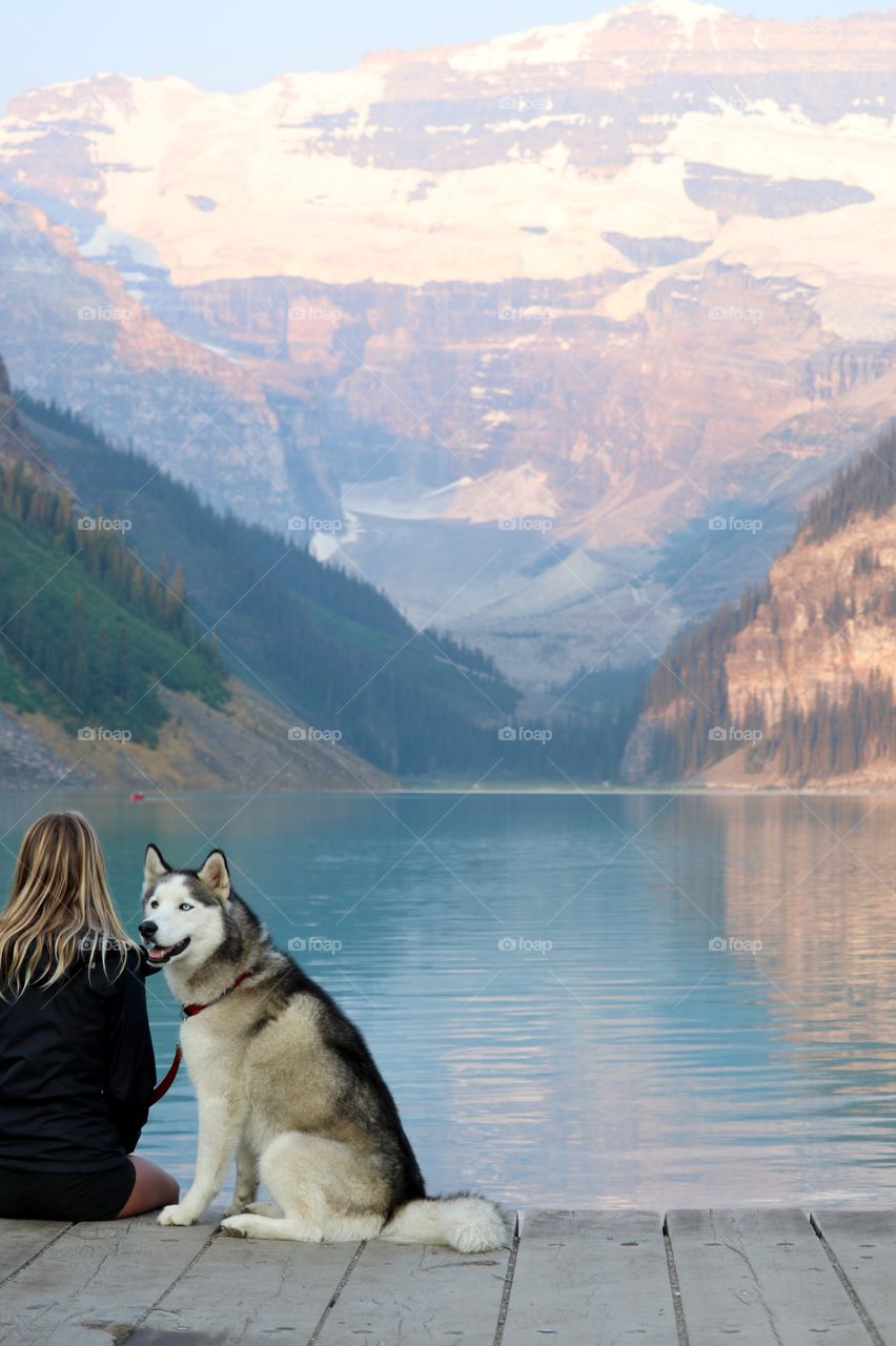 Blonde haired woman sitting beside beautiful scenic glacial Lake Louise in the Rocky Mountains of Alberta Canada near Banff, with blue eyed husky dog, 