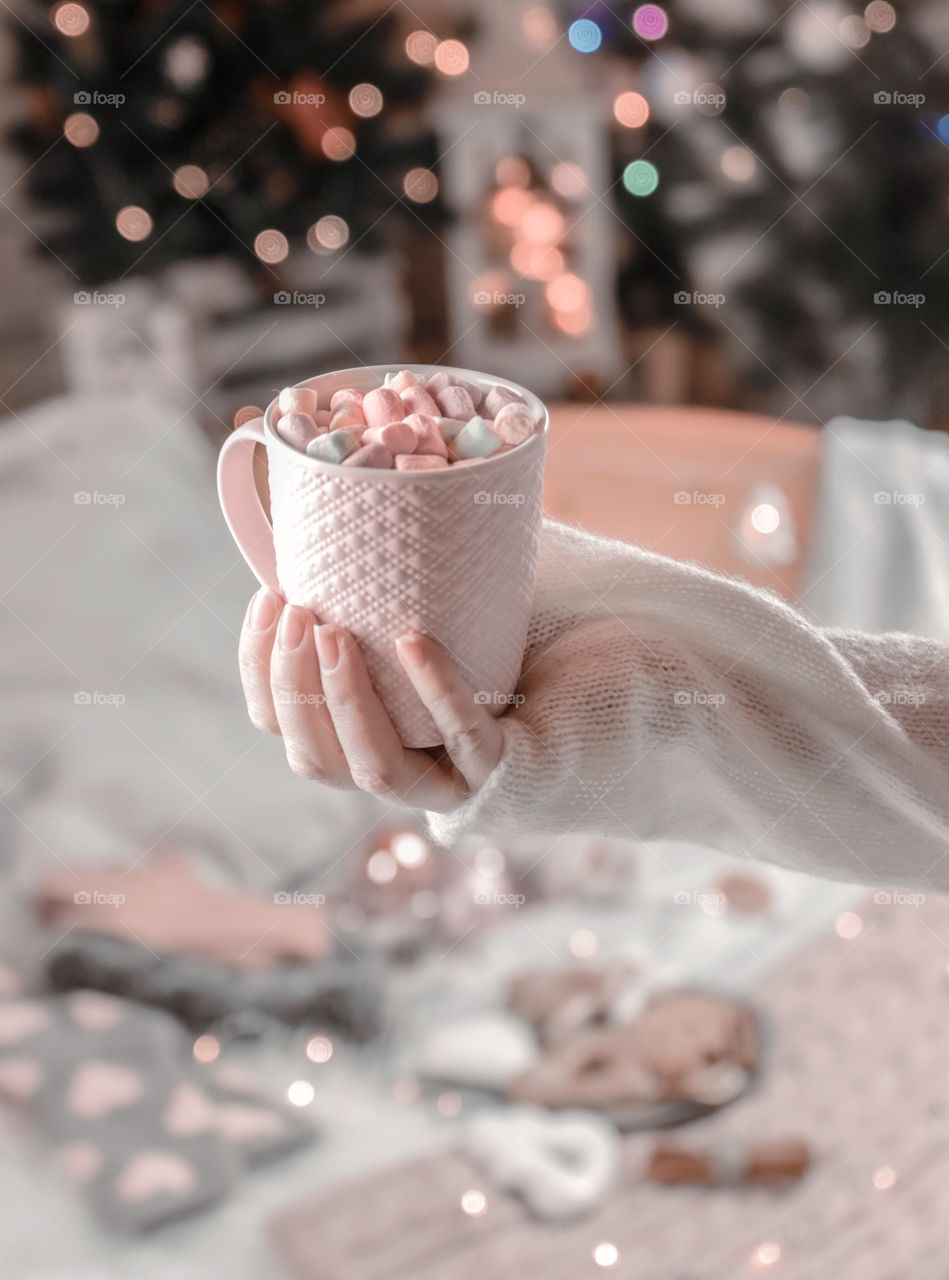  
Cup with marshmallows in female hands on a background of Christmas decor