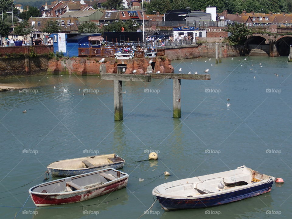 Boats in the foreground of Folkestone Harbor in England with birds on posts and people enjoying food and drink on a brick deck on a sunny summer day. 