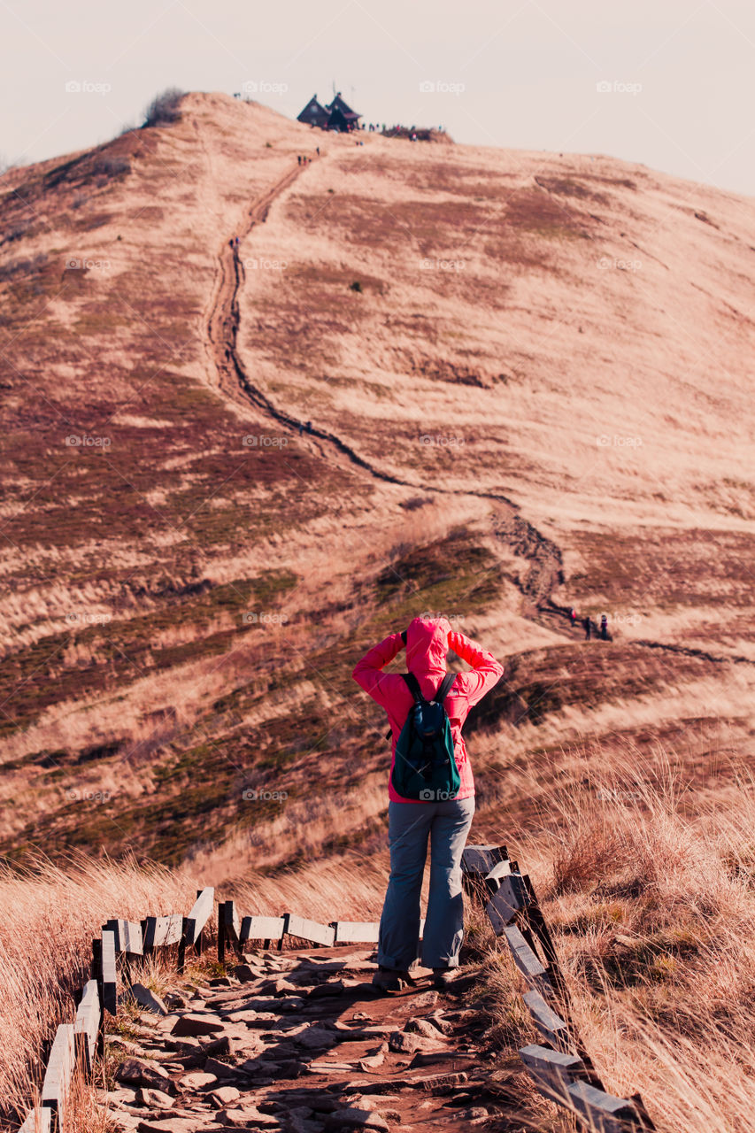 Woman walking along mountain path admiring mountains scenery during a trip on sunny windy autumn day. Hiker wearing sports clothes and shoes carrying backpack on her back. There is grassy hillside coloured with browns around footpath in fall season