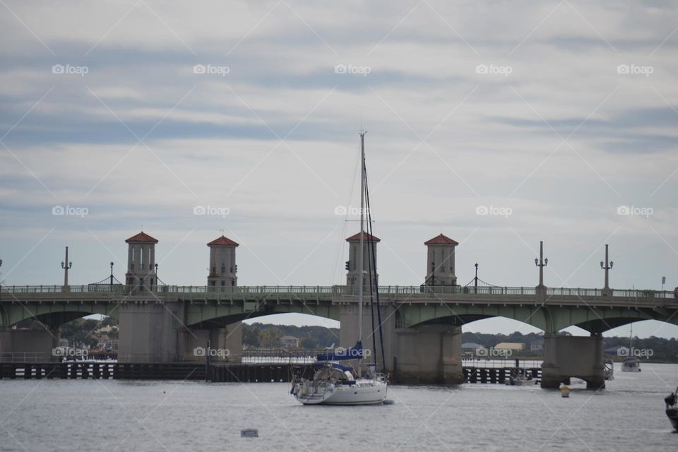 A concrete draw bridge across gray water with a sailboat in front of it and a cloudy blue sky behind