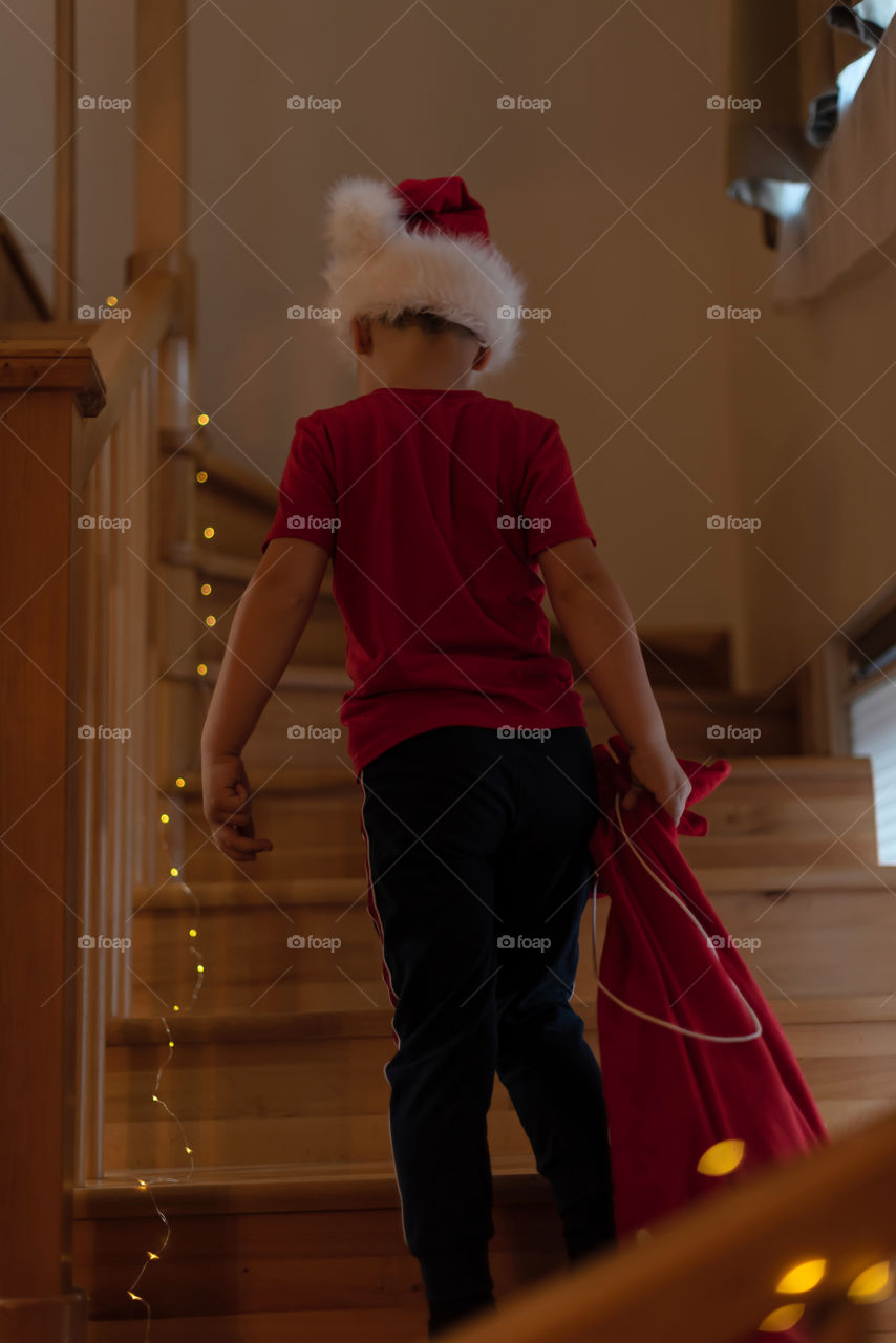 Little boy in Santa hat carries a bag of gifts up the stairs. Christmas or Happy New Year concept. Xmas spirit