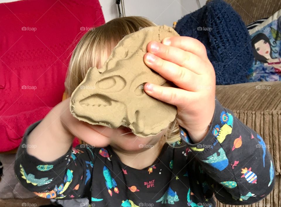 Close up of a child’s hand, holding onto the head of a sand sculptured triceratops dinosaur. Whilst wearing a dinosaur themed top. 