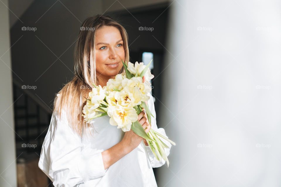 Young beautiful smiling woman forty year with blonde long hair in white shirt with bouquet of yellow flowers in hands near window in bright interior at the home