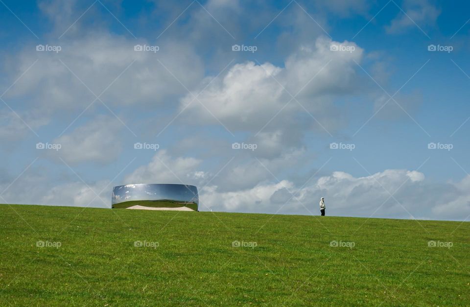 At the top of a grassy hill outside Brighton, Anish Kapoor’s C-Curve (2007) is exhibited, reflecting the blue and cloudy sky in its distorted mirrors. A lone spectator is admiring the scenery