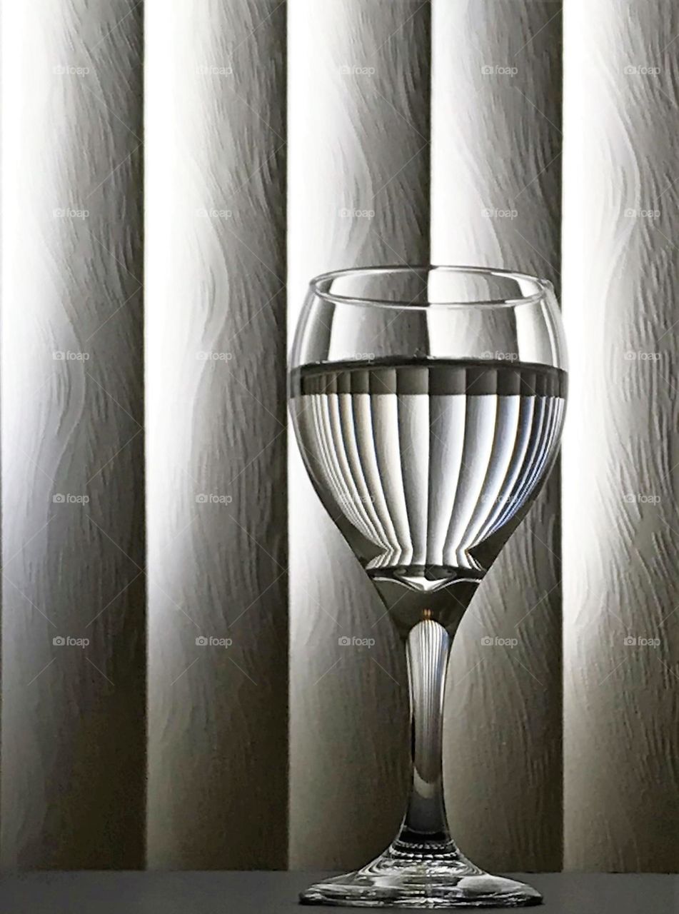 Black and white still life of wine glass partially filled with water against a window blind