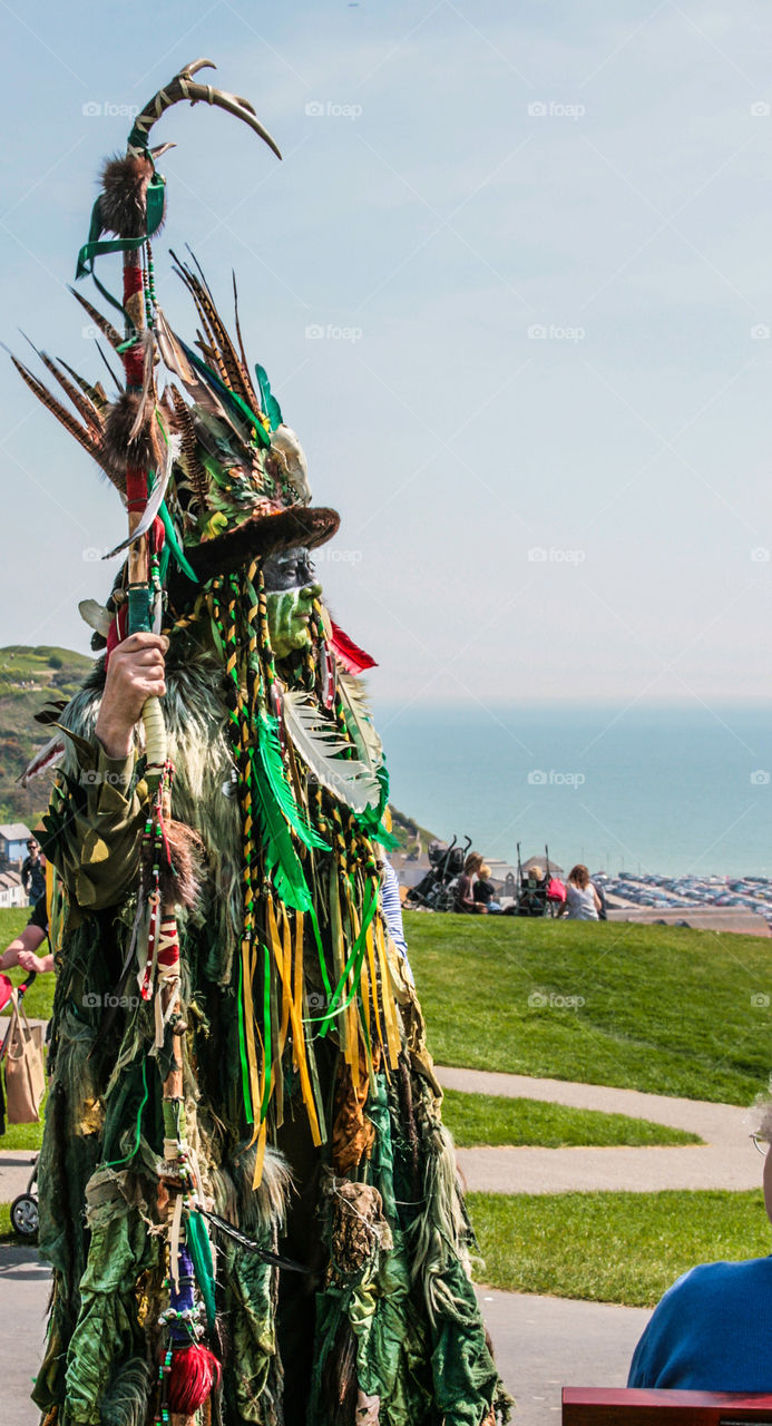 A man dressed up in leaves and feathers and greenery, holding a staff, with the sea in the distance at Hastings Traditional Jack in the Green, U.K. 2008