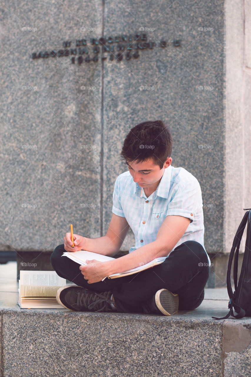 Student making the notes learning from books sitting on a monument outside of university. Young boy wearing a blue shirt and dark jeans