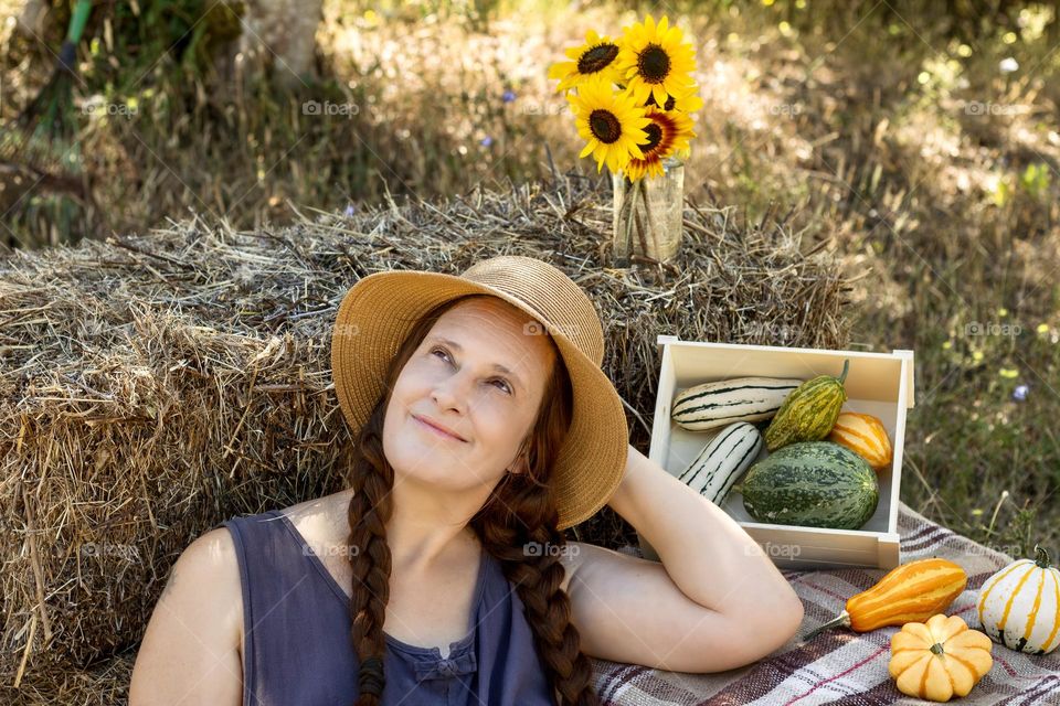 Woman relaxes in late summer on hay bales with sunflowers and autumn gourds. 